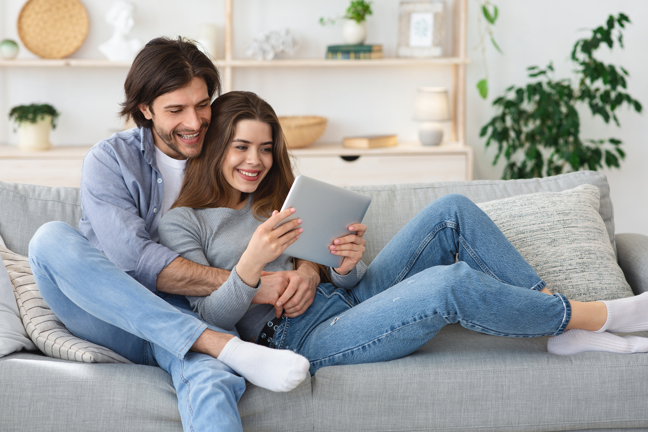 Cheerful couple cuddling on sofa at home, using digital tablet