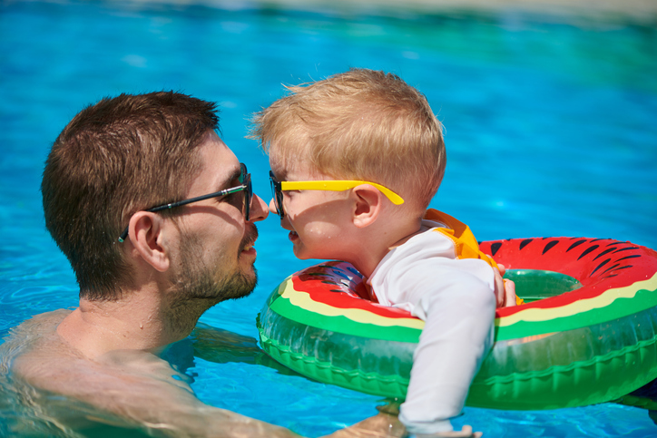 Toddler boy floating in a watermelon shape floatie in swimming pool touching noses with father and both smiling. Charleston Wesley Chapel