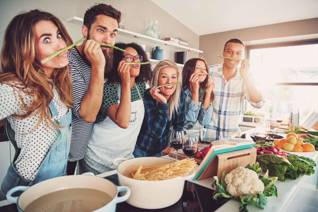 group of friends making silly faces and holding asparagus above there smiles, drinking wine and making pasta in a kitchen