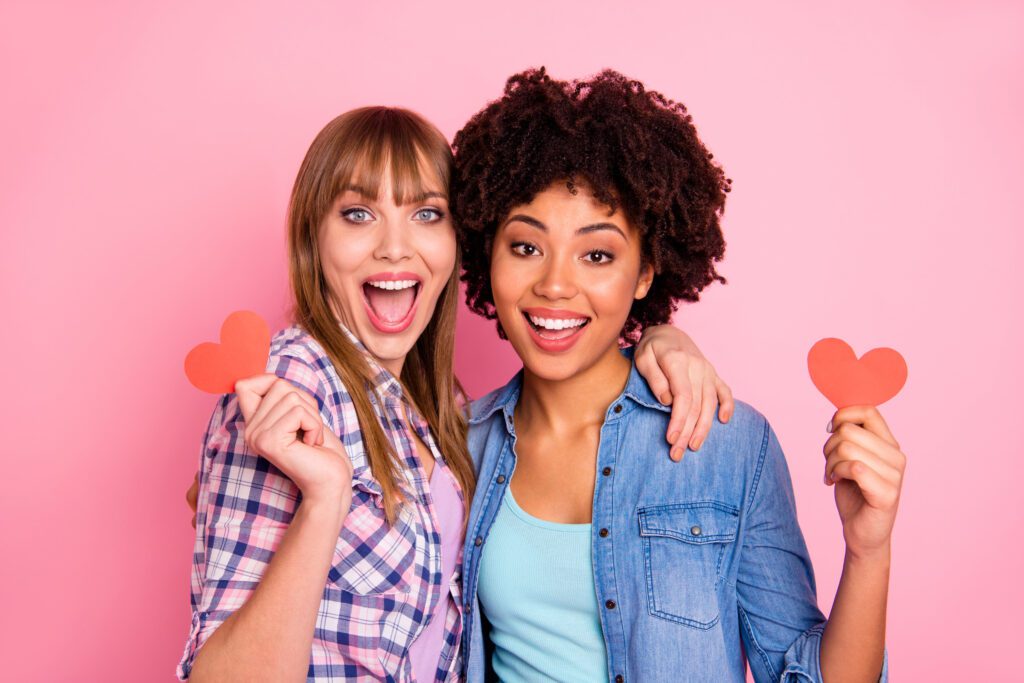 Close-up portrait of two person nice cute lovely sweet attractive charming cheerful girls in checkered shirt holding in hands small little cards cuddling isolated over pink pastel background.